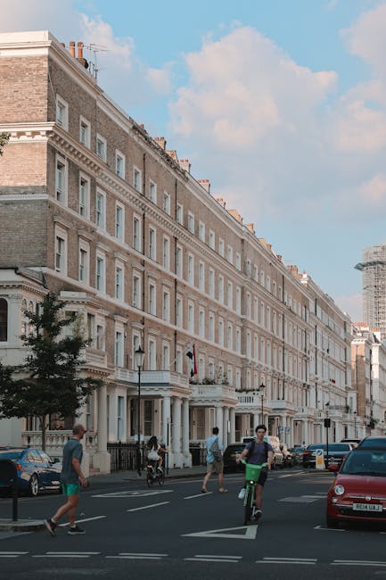 This image shows a row of elegant, cream-colored Georgian-style terraced houses with detailed masonry and large windows, located on a busy street in Kensington. The foreground features pedestrians wal