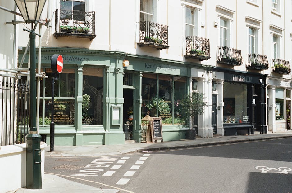 Exterior view of a row of shops in Kensington, London, with a light green and black storefront. The building features large glass windows displaying plants and decor inside, framed by white trim. Above, there are several small balconies with wrought iron railings and potted plants. A sidewalk with a pedestrian crossing is visible, along with street signs and a bicycle lane marking on the road. The scene is well-lit with natural daylight, presenting a clean and inviting urban shopping street, highlighting the area where Carpet Cleaners Kensington offers professional cleaning services near Palace and in Kensington.