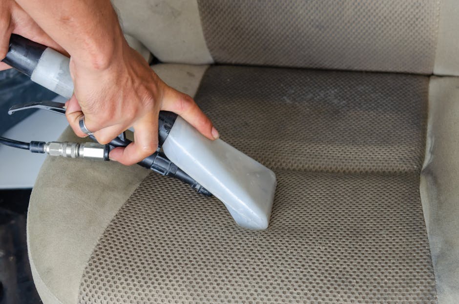 A close-up of a person's hand holding a professional upholstery cleaning wand, being used to deep clean a beige fabric car seat or soft furniture. The wand is connected to a hose and appears to be emitting a spray or suction, indicating a thorough surface cleaning process. The fabric seat has a textured, dotted pattern, with visible dirt and stains being targeted for removal. The surrounding upholstery is clean and well-maintained, demonstrating effective interior cleaning techniques. Natural light highlights the fabric’s texture and cleanliness, and the setting suggests a focus on fabric or furniture surface sanitisation by Carpet Cleaners Kensington, reflecting their expertise in domestic cleaning and upholstery surface cleaning near Kensington Palace.