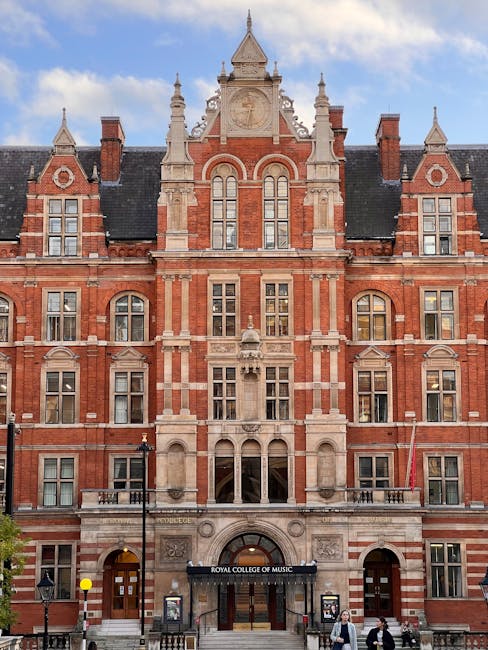 A detailed exterior view of the Royal College of Music building in Kensington, featuring red brick and stone architectural elements, large arched windows, decorative spires, and intricate stone carvin
