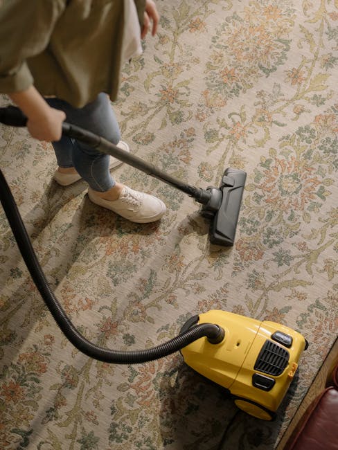 A person using a yellow vacuum cleaner with a black hose attachment to deep clean a patterned, beige and pastel-colored rug in a domestic setting. The individual is wearing white shoes and beige pants, and is operating the vacuum on the intricate floral design of the carpet. The room has soft, natural lighting, highlighting the clean, dust-free surface, and the vacuum is positioned near a piece of furniture with a wooden finish. This scene demonstrates surface cleaning and maintenance for a residential space, with professional insights from Carpet Cleaners Kensington.
