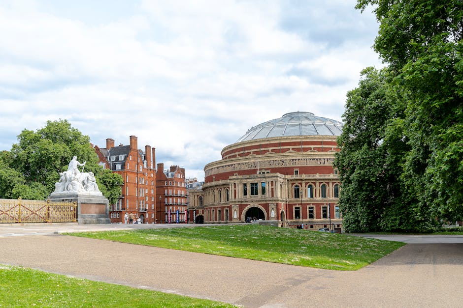 A historic circular building with a domed roof, featuring brick and stone architectural details, is situated in a park surrounded by lush green trees. The area includes a pathway, well-maintained grass, and a monument sculpture on the left side, set against a partly cloudy sky. The scene depicts a peaceful, outdoor setting in Kensington, with natural light highlighting the building's features, and emphasizing the cleanliness and well-kept landscape as part of the area’s overall hygiene and maintenance appearance, aligning with surface cleaning and sanitisation themes. Carpet Cleaners Kensington is not visible in this image.