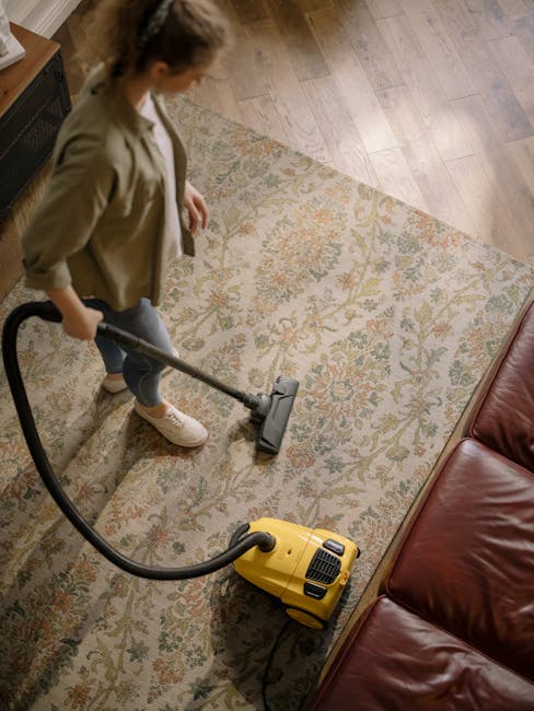 A person using a yellow vacuum cleaner with a black hose attachment to deep clean a patterned, beige and pastel-colored rug in a domestic setting. The individual is wearing white shoes and beige pants, and is operating the vacuum on the intricate floral design of the carpet. The room has soft, natural lighting, highlighting the clean, dust-free surface, and the vacuum is positioned near a piece of furniture with a wooden finish. This scene demonstrates surface cleaning and maintenance for a residential space, with professional insights from Carpet Cleaners Kensington.