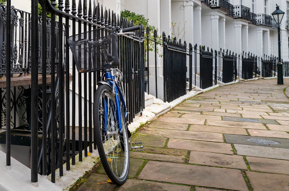 A blue bicycle leaning against a black wrought iron fence alongside a white residential building. The sidewalk is paved with large, rectangular stone slabs, some with visible moss or dirt in the joints. The building features white columns, balustrades, and black iron railings on the steps, with potted plants and greenery visible near the entrance. Bright natural lighting highlights the cleanliness of the surfaces, and the scene is typical of a tidy, well-maintained Kensington street. Carpet Cleaners Kensington specializes in surface cleaning and deep cleaning services to support hygienic and well-maintained residential environments, as referenced in their page on Kensington council waste rules for mattress and sofa disposal.
