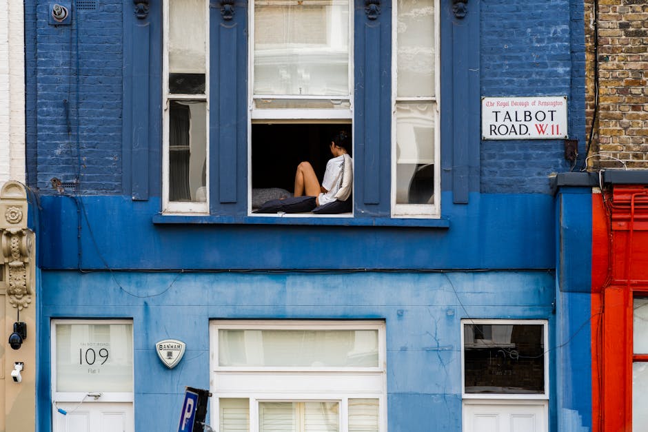 A young woman sits on a windowsill of a blue-painted building in Kensington, with her legs drawn up and her back leaning against the window frame. The window is open, revealing a glimpse of an interior space, and the room appears to be illuminated by natural daylight. The building's façade includes decorative wooden trim around the window and a white door below with a small window and a security alarm sign. Adjacent brickwork and a street sign indicating Talbot Road W11 are visible, with a clear, bright outdoor setting. The scene emphasizes the architectural details and the contrast of colors between the building's vibrant blue paint and the neighboring structures, captured in a neutral, documentary-style composition.