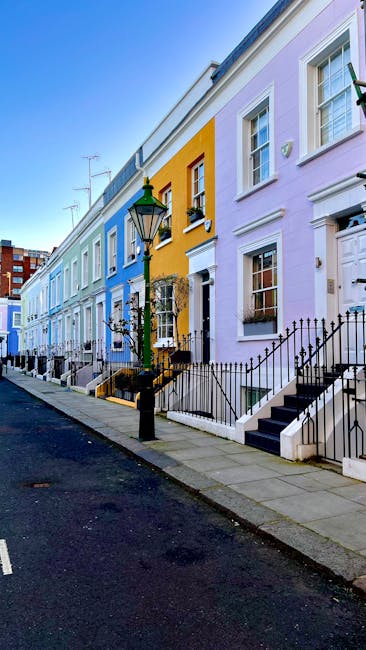 A row of colorful terraced houses in a residential street, featuring pastel pink, yellow, and blue facades with white window frames and black iron railings. The pavement is clean and well-maintained, 
