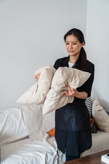 Close-up of a person using a handheld vacuum cleaner to deep clean the fabric upholstery of a green, button-tufted armchair in a living room. The chair's surface appears slightly textured, with visibl